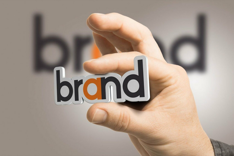 A hand holding a cut-out of the word brand with a blurred background of the same word.