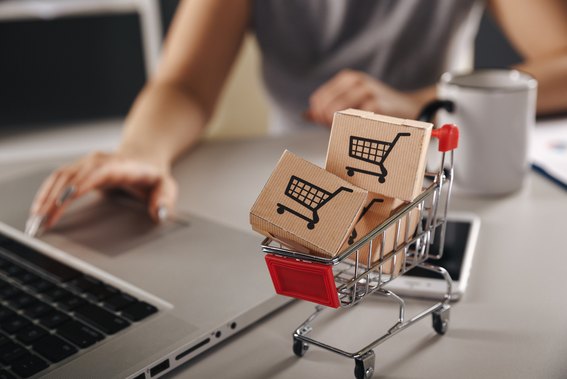 A person is using a laptop with a miniature shopping cart filled with small boxes nearby.